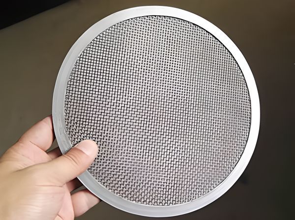 A hand is holding a piece of galvanized woven wire mesh filter disc.